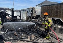 Firefighter in turnout gear sprays water over smoldering debris from a burned structure; a trailer sits in the background.