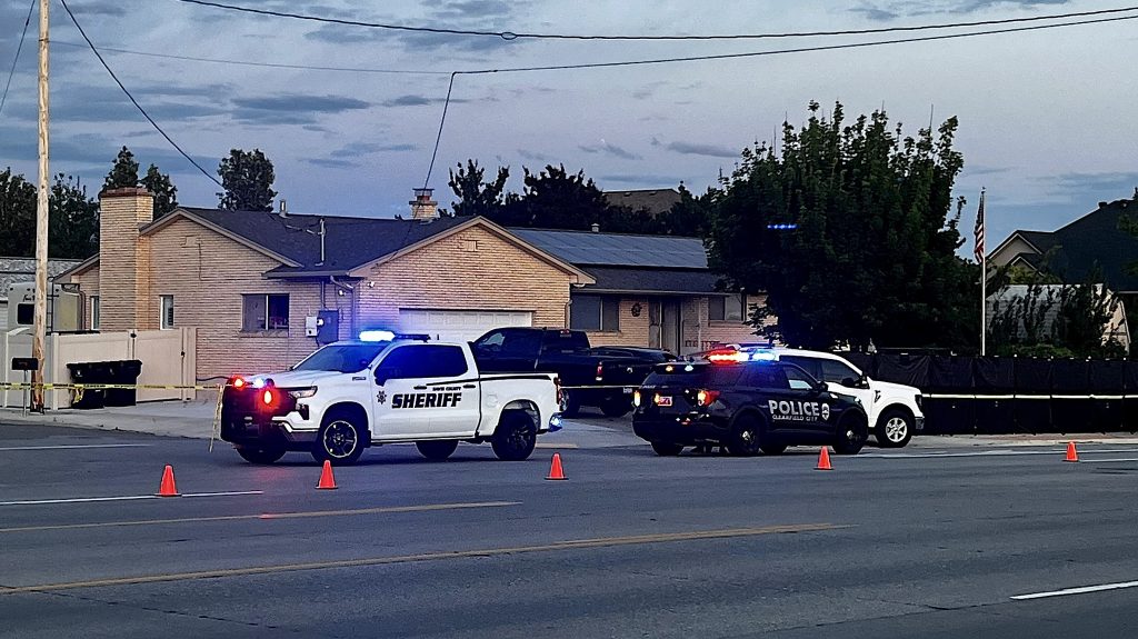 Police and sheriff cars with flashing lights blocking a residential street; yellow crime tape and orange cones mark the area.