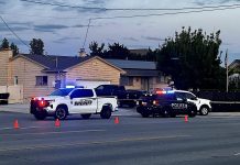 Police and sheriff vehicles with flashing blue lights block a suburban street; orange cones and caution tape mark off a scene in front of houses.]