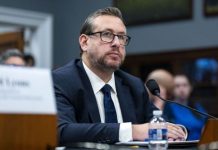 Man in a dark suit and glasses seated at a hearing table with a microphone and water bottle nearby.