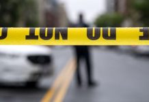Yellow caution tape reading 'DO NOT ENTER' stretches across a street, with a blurred figure and police car in the background.