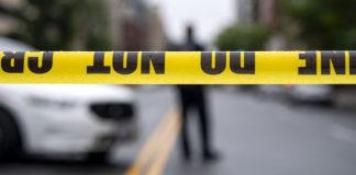 Yellow caution tape reading 'DO NOT ENTER' stretches across a street, with a blurred figure and police car in the background.