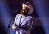 Bearded man in a white cowboy hat and patterned blue jacket singing into a microphone on stage with spotlights.