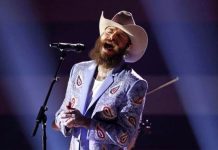 Bearded man in a white cowboy hat and patterned blue jacket singing into a microphone on stage with spotlights.
