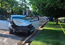 Car crash on a street with the front end heavily damaged and both doors open; yellow caution tape and responders in the background, suggesting an accident scene.