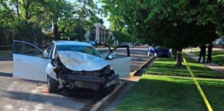 Car crash on a street with the front end heavily damaged and both doors open; yellow caution tape and responders in the background, suggesting an accident scene.