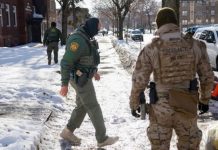 Two tactical officers in winter gear patrol a snow-covered residential street, with parked cars and bare trees nearby.