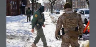 Two tactical officers in winter gear patrol a snow-covered residential street, with parked cars and bare trees nearby.