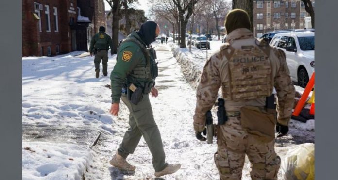 Two tactical officers in winter gear patrol a snow-covered residential street, with parked cars and bare trees nearby.