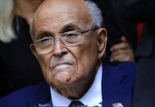 Older man with glasses in a blue suit, frowning slightly and looking to the side.