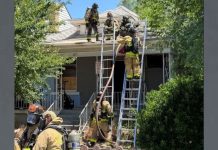 Firefighters on ladders at the roof peak of a suburban home during an emergency response.