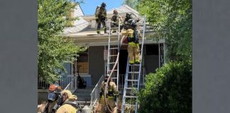 Firefighters on ladders at the roof peak of a suburban home during an emergency response.