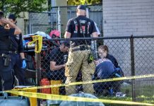 Outdoor crime scene with police and investigators behind a chain-link fence, yellow caution tape, and equipment bags nearby.