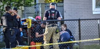 Outdoor crime scene with police and investigators behind a chain-link fence, yellow caution tape, and equipment bags nearby.