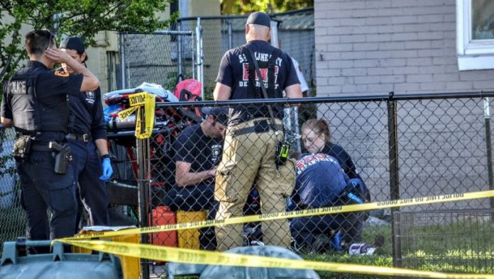 SLC Romy 5 Outdoor crime scene with police and investigators behind a chain-link fence, yellow caution tape, and equipment bags nearby.