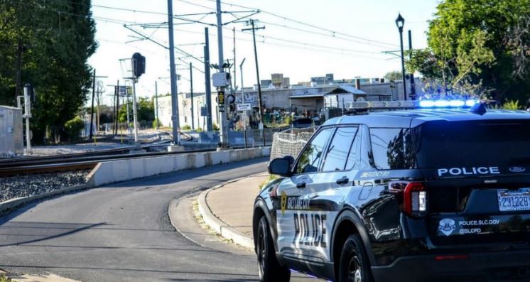 Black and white police SUV with flashing blue lights at a street intersection near railroad tracks, suggesting a police presence.