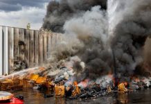 Industrial fire: thick black smoke rising from burning pallets and debris beside a fence, with firefighters spraying water on the blaze. (informative)