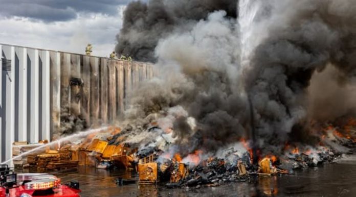 SLCFD 3 Industrial fire: thick black smoke rising from burning pallets and debris beside a fence, with firefighters spraying water on the blaze. (informative)