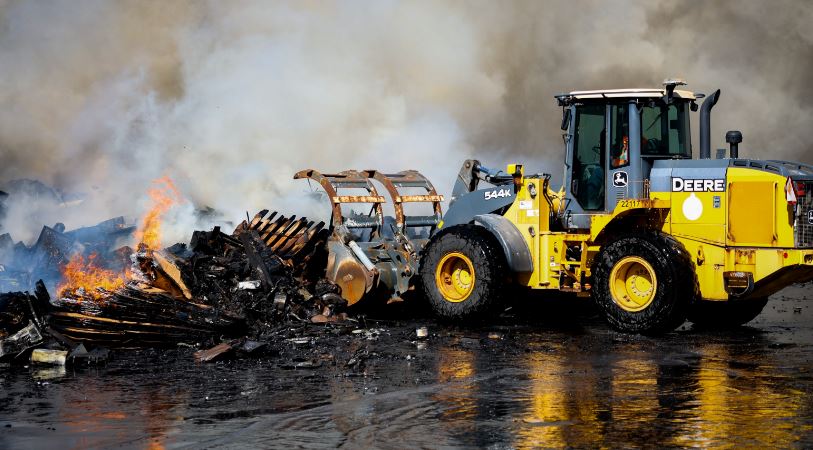Yellow John Deere heavy loader moving a pile of charred debris as flames and smoke rise in the background on a wet surface