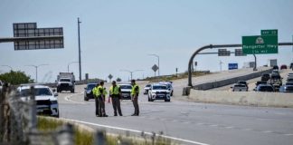Four police officers in neon vests stand together on a highway shoulder while cars pass by and a checkpoint is set up ahead on a divided freeway near an Exit 13 sign labeled Bangerter Hwy.