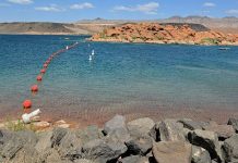 Calm lake with a curve of red buoys leading to a rocky shoreline and distant desert hills under a blue sky.