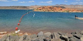 Calm lake with a curve of red buoys leading to a rocky shoreline and distant desert hills under a blue sky.