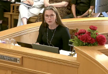 Woman at a wooden dais speaking into a microphone; nameplate reads 'Mayor' with red flowers on the side.