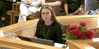 Woman at a wooden dais speaking into a microphone; nameplate reads 'Mayor' with red flowers on the side.