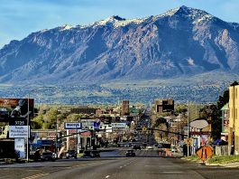 Wide city street with cars and billboards, mountains in the distance under a blue sky.