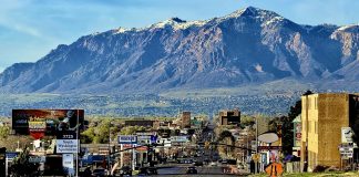 Wide city street with cars and billboards, mountains in the distance under a blue sky.
