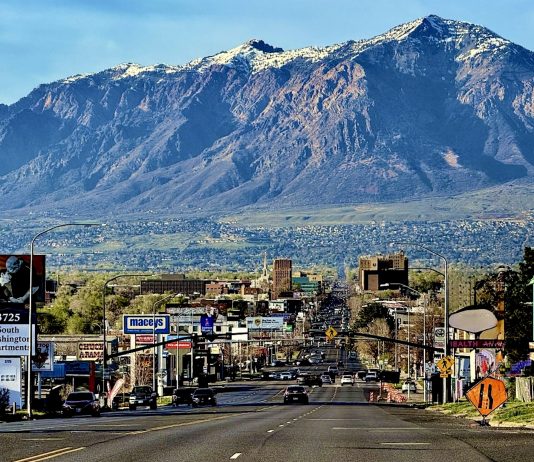 Wide city street with cars and billboards, mountains in the distance under a blue sky.