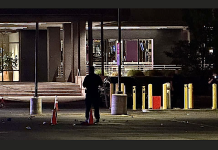 Nighttime exterior of a building with a silhouetted person near orange cones and yellow bollards in a dim parking lot nearby storefronts.”,