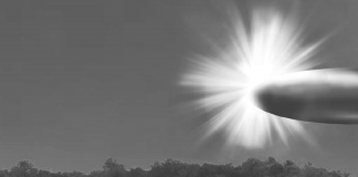 Grayscale landscape showing a bright sunburst on the right, rays radiating across the sky over a tree line and open field; a small rectangular object sits in the foreground left of center.