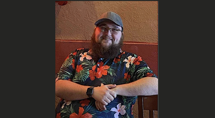 Unsolved 2023 Springville Homicide Smiling man with beard and glasses wearing a colorful Hawaiian shirt and a gray cap, seated with arms folded on a chair.