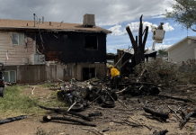 Fire-damaged two-story house with a charred exterior, scattered debris, and a firefighter in a yellow uniform examining the rubble amid burnt trees.