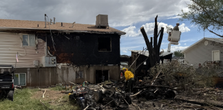 Fire-damaged two-story house with a charred exterior, scattered debris, and a firefighter in a yellow uniform examining the rubble amid burnt trees.