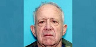 Mugshot-style portrait of an older man with gray hair and a stern expression against a blue background.