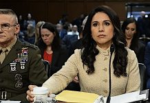 Tulsi Gabbard resigns as director of national intelligence, cites husband’s cancer diagnosis Woman in a beige coat testifying at a formal hearing, seated at a table with a microphone and papers, surrounded by attendees and officials.