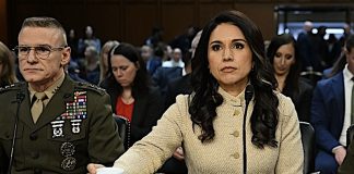 Woman in a beige coat testifying at a formal hearing, seated at a table with a microphone and papers, surrounded by attendees and officials.
