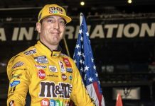 Smiling race car driver in a yellow sponsor-filled suit and cap stands with an American flag at a motorsports venue.