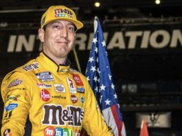 Smiling race car driver in a yellow sponsor-filled suit and cap stands with an American flag at a motorsports venue.