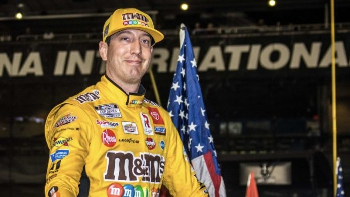 Smiling race car driver in a yellow sponsor-filled suit and cap stands with an American flag at a motorsports venue.