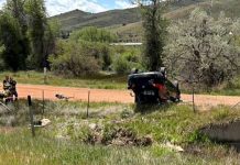 SUV flipped upside down on a dirt road in a rural area, with two responders nearby and hills in the background.
