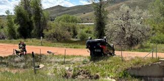 SUV flipped upside down on a dirt road in a rural area, with two responders nearby and hills in the background.