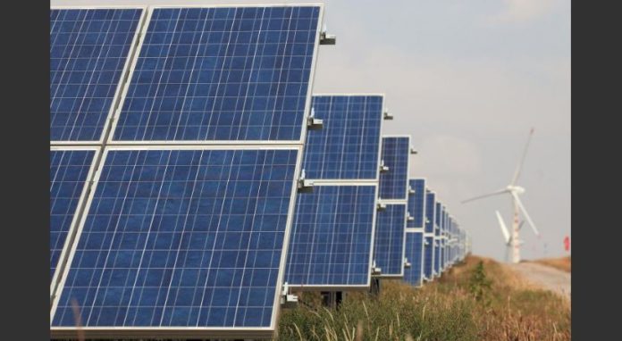 Row of blue solar panels in a field with a wind turbine visible in the distance.