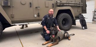 K-9 police dog in a tactical vest lying on the driveway beside a uniformed officer in front of a large armored vehicle.