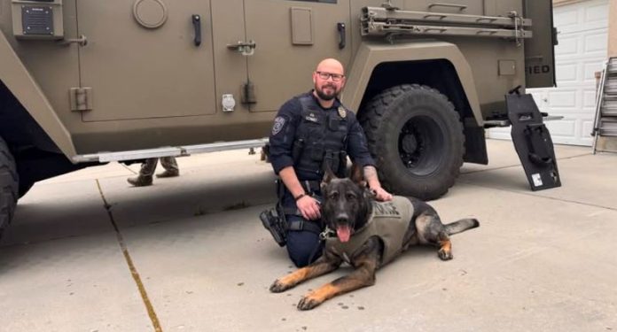 K-9 police dog in a tactical vest lying on the driveway beside a uniformed officer in front of a large armored vehicle.