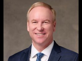 Smiling man in a navy suit, white shirt, and blue patterned tie, against a gray backdrop (professional headshot).