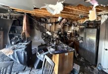 Charred kitchen with a collapsed ceiling and burnt debris strewn across a table and chairs after a house fire, showing extensive damage outside of frame edges.