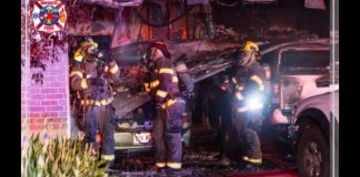 Firefighters in full gear work at a car fire outside a brick building, with a damaged SUV and a hose on the ground outside.